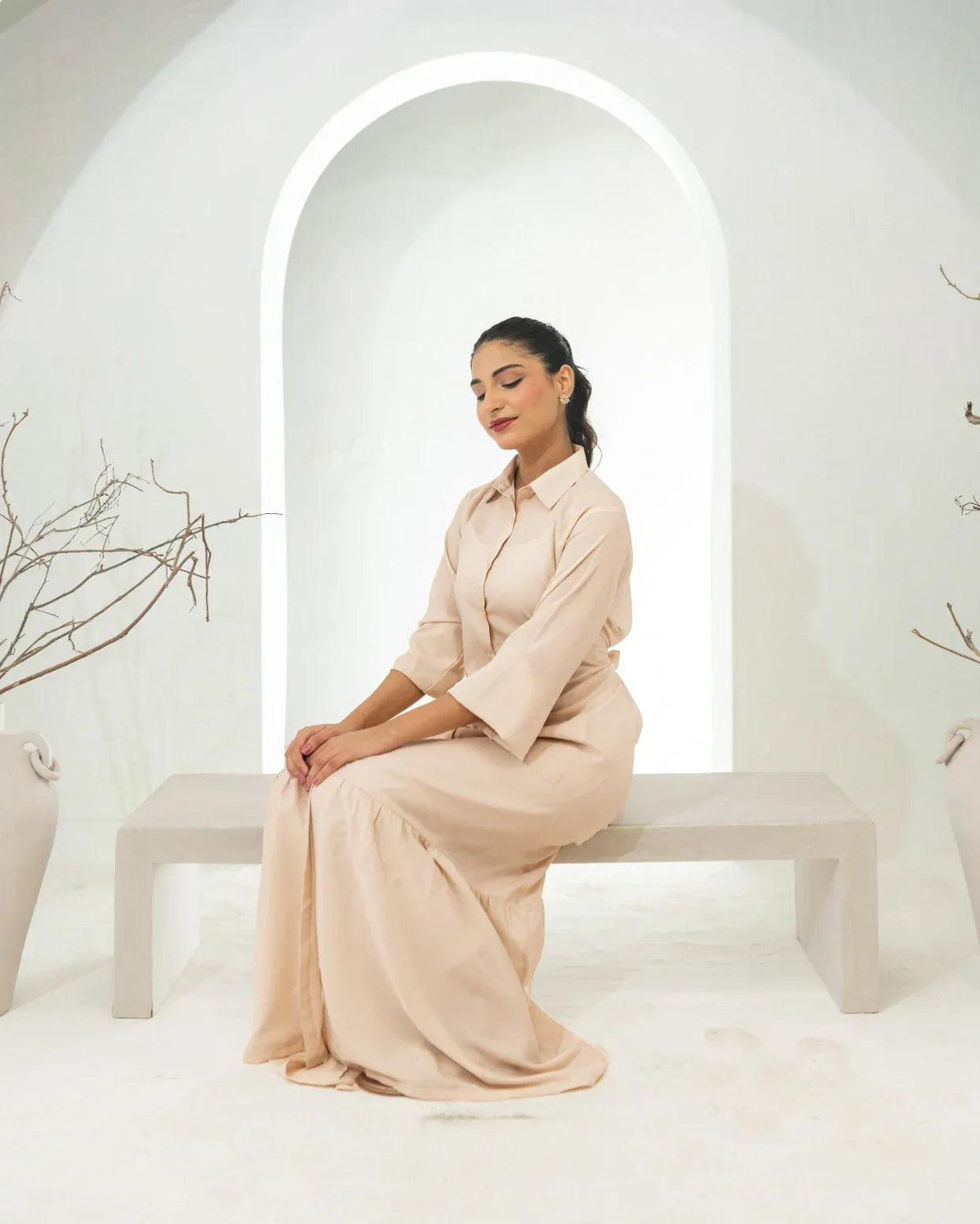 Woman in a fawn outfit sitting on a white bench in a minimalistic setting with arches and plants.