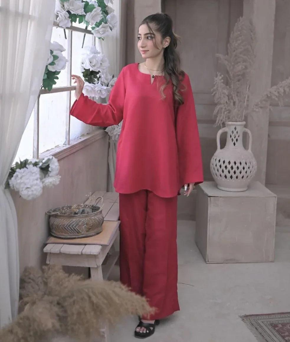 A woman in a deep pink outfit standing against a window, She has a calm expression and wears black sandals.