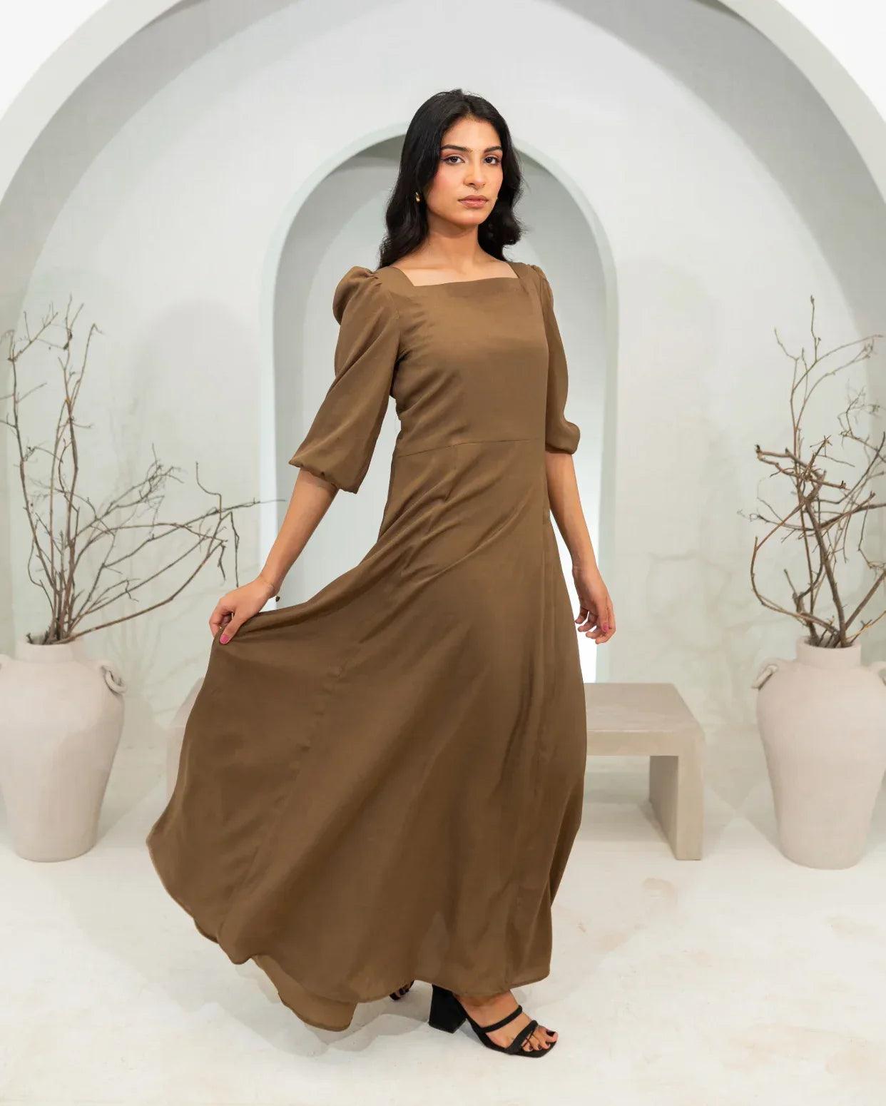 Woman wearing a khaki color long dress standing in a minimalistic setting with white arches and potted plants.