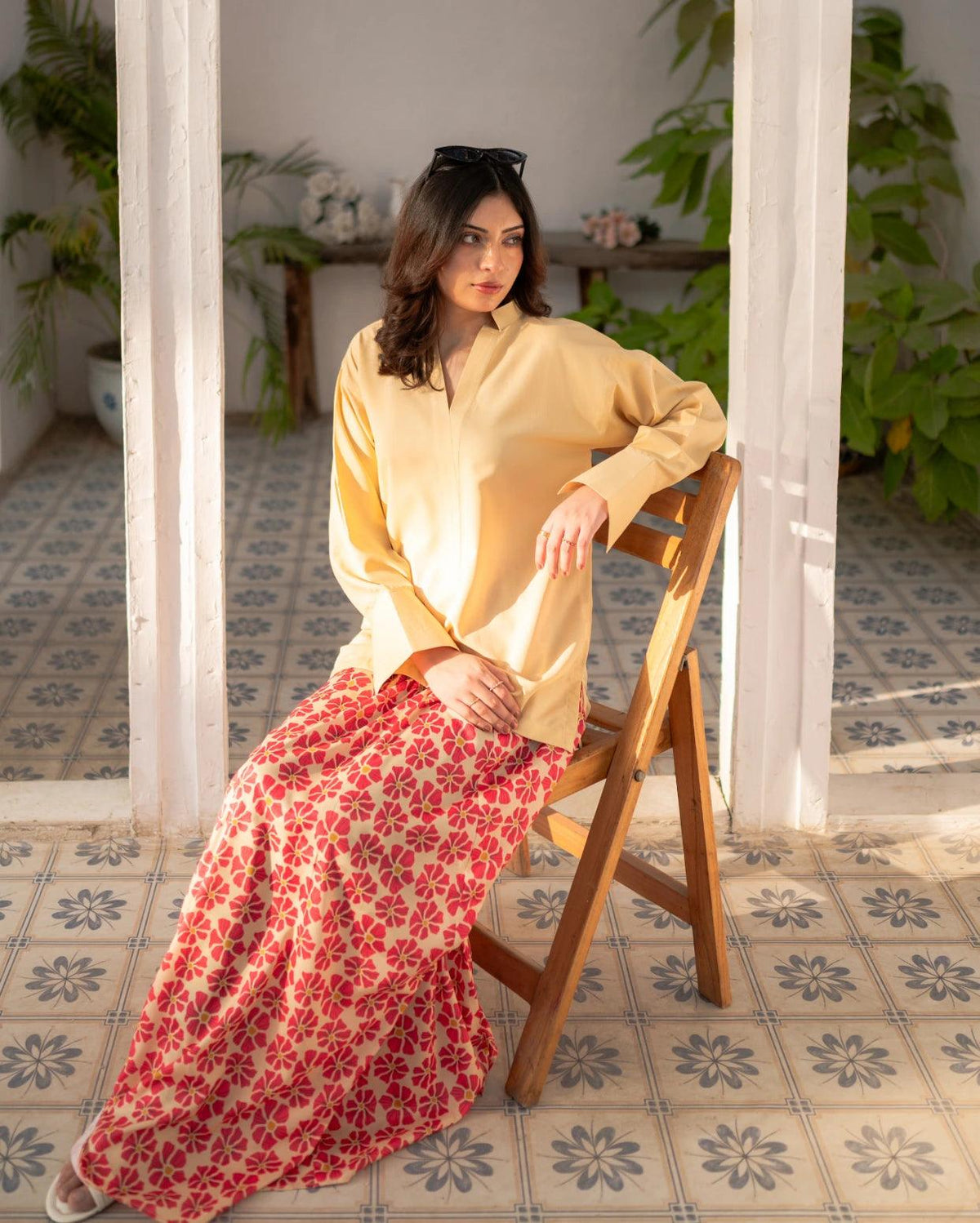 Woman sitting on a wooden chair wearing a lemon yellow top and red floral skirt in a room with plants and patterned floor.