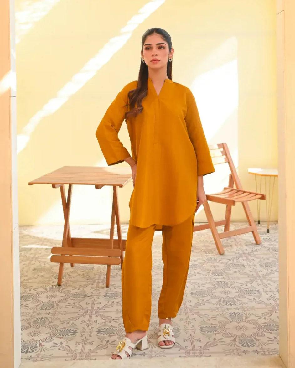 A woman wearing a mustard yellow two-piece dress with long sleeves, consisting of a shirt and trousers, standing in a sunny area next to a wooden bench.