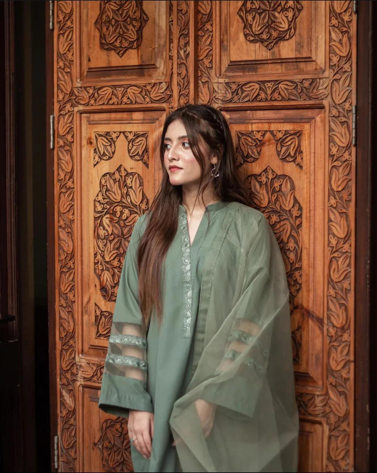 A woman in an olive green stitched dress stands confidently against an ornately carved wooden door, conveying elegance and cultural richness.