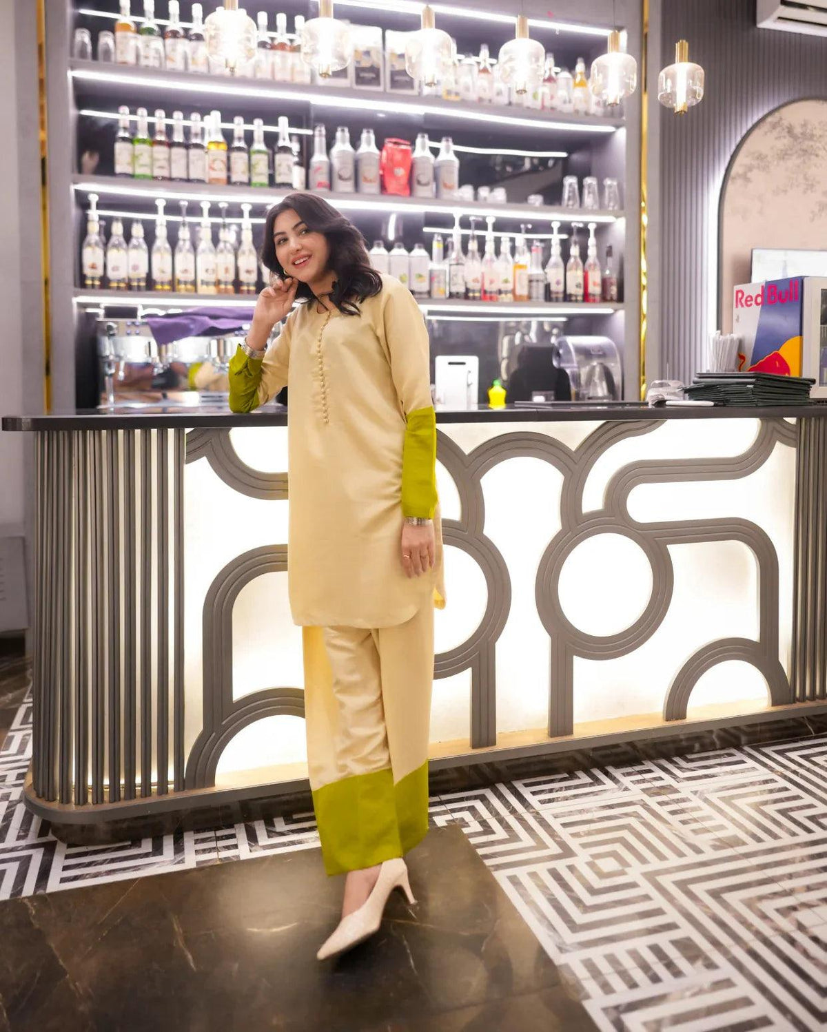 Woman in a beige and green outfit standing in front of a bar counter with drinks.