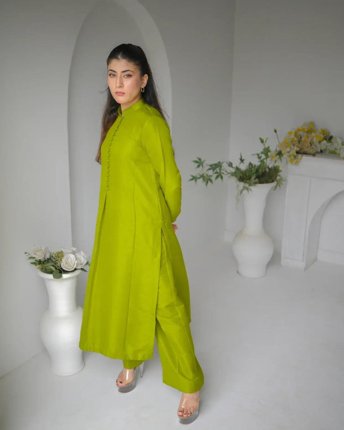 Woman wearing a mehndi/green dress standing in a minimalistic room with white walls and decorative vases.