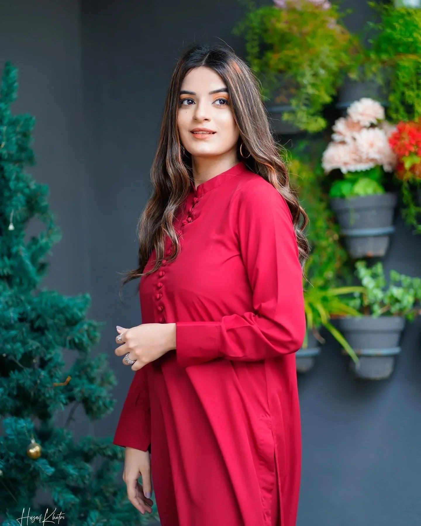 Woman in a red dress stands confidently near a small decorated tree, with a backdrop of green plants on a gray wall, conveying elegance.