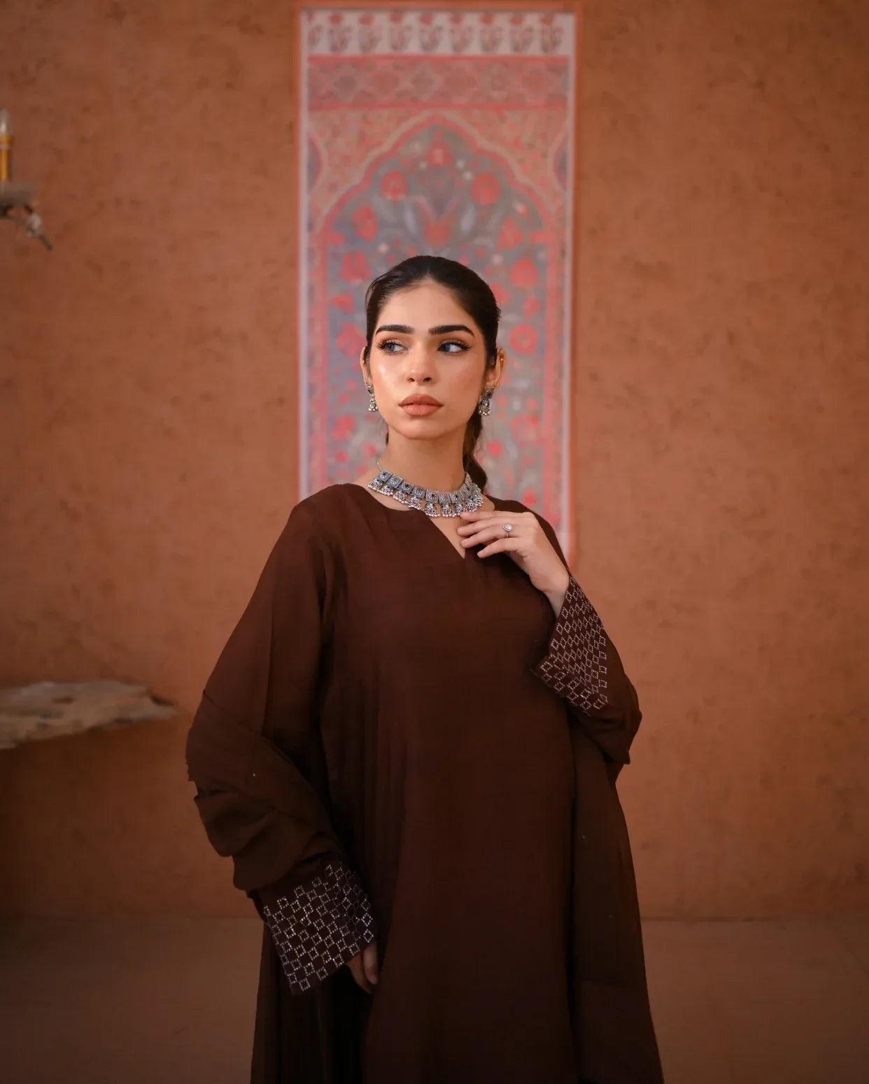 A woman wearing a brown rawsilk dress which includes a shirt and a chiffon dupatta. The dress is embellished with stone details.