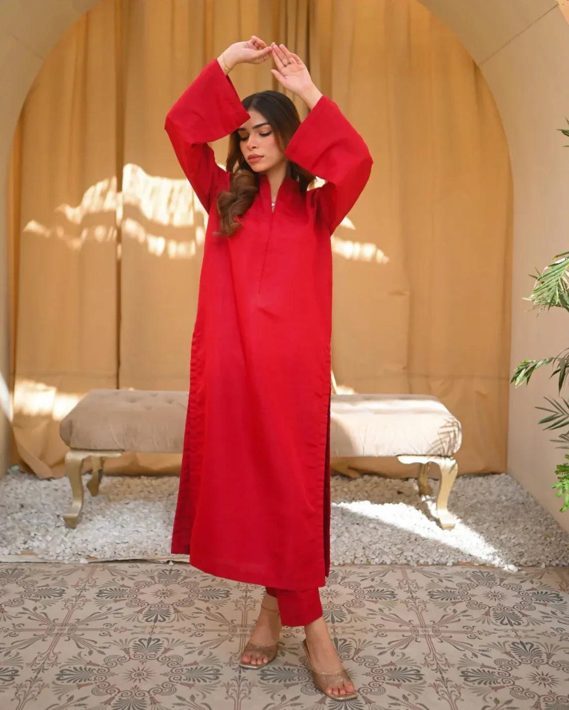 A woman standing in a sunny room wearing a red two-piece rawsilk dress with a long shirt and straight trousers, posing next to a beige bench and a green plant.
