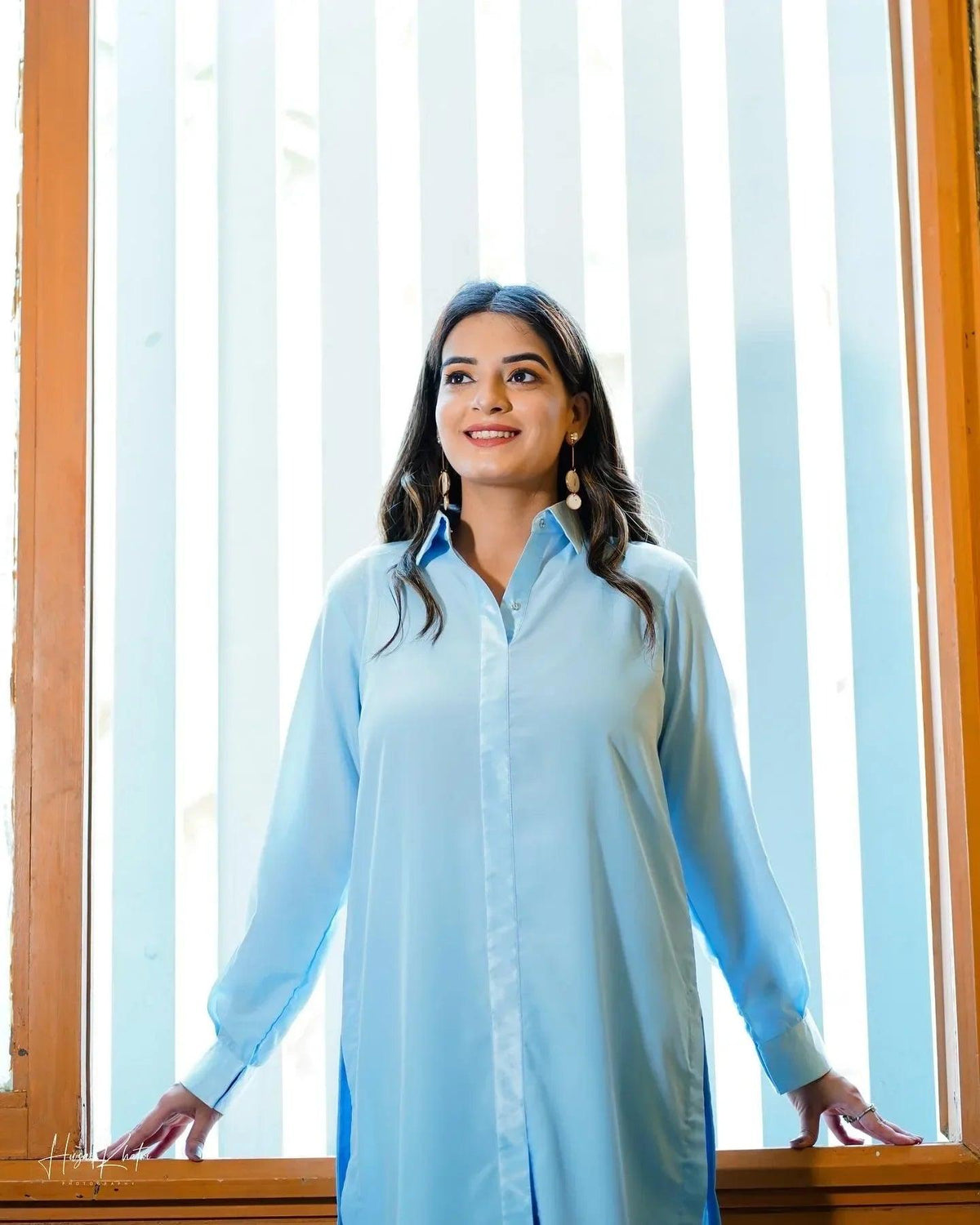 A woman in a sky blue traditional dress poses confidently indoors. She stands near a window