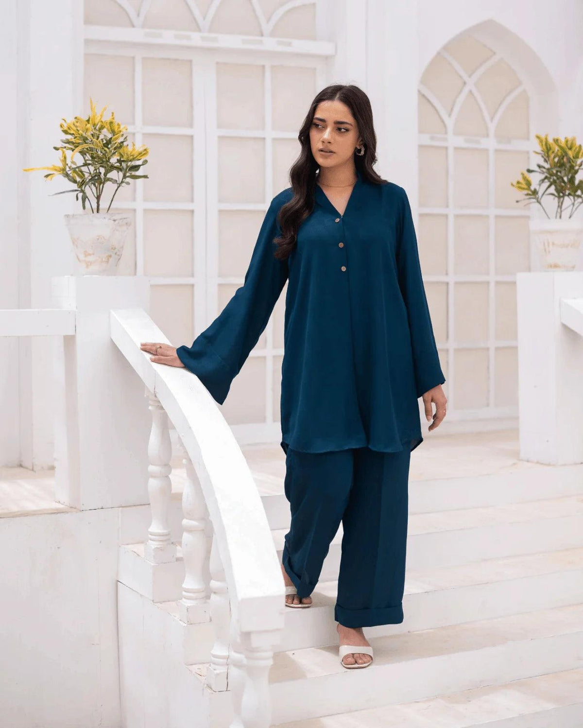 A woman wearing a teal blue co ord set with wide sleeves, standing on white stairs in a bright, white architectural setting.