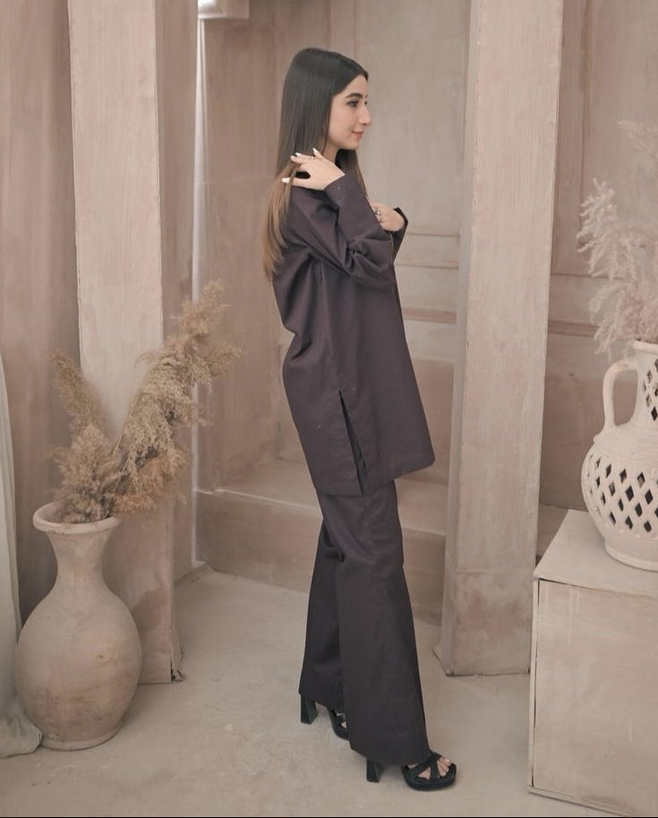 A woman in a stylish dark brown outfit stands in a minimalist room with decorative vases and pampas grass. The ambiance is calm and elegant.
