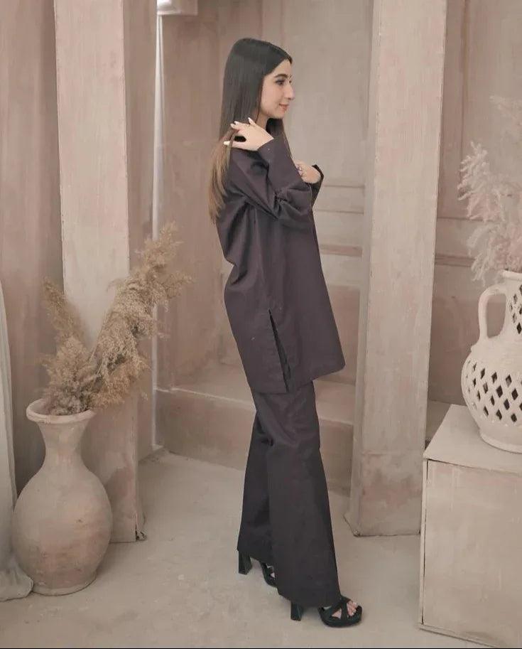 A woman in a stylish dark brown outfit stands in a minimalist room with decorative vases and pampas grass. The ambiance is calm and elegant.