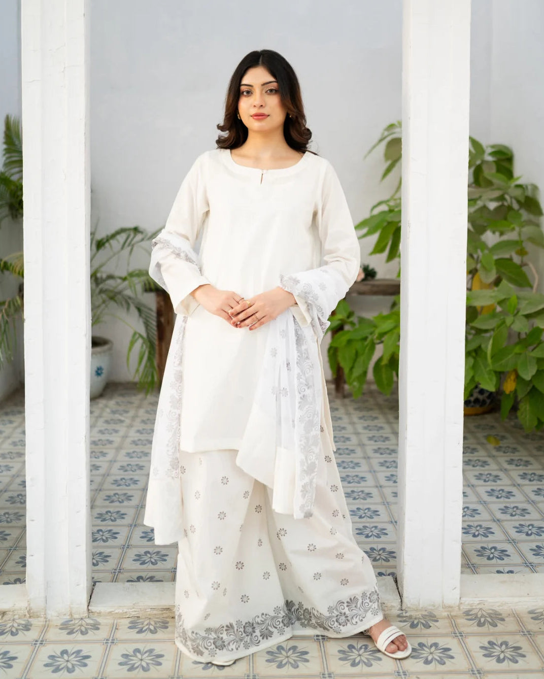 Woman in a white traditional kurta set standing in a room with plants and patterned floor.