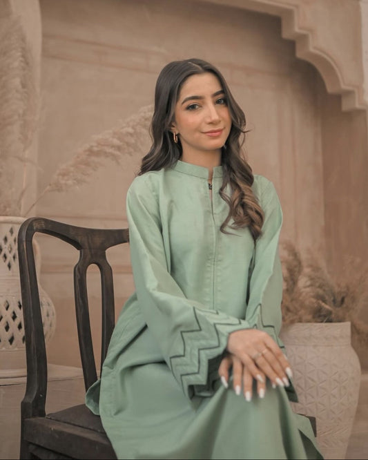 A Woman wearing an apple green dress with embroidery on sleeves and zigzag cuffs, smiling confidently. Neutral-toned vases with dried plants enhance the serene, stylish setting.