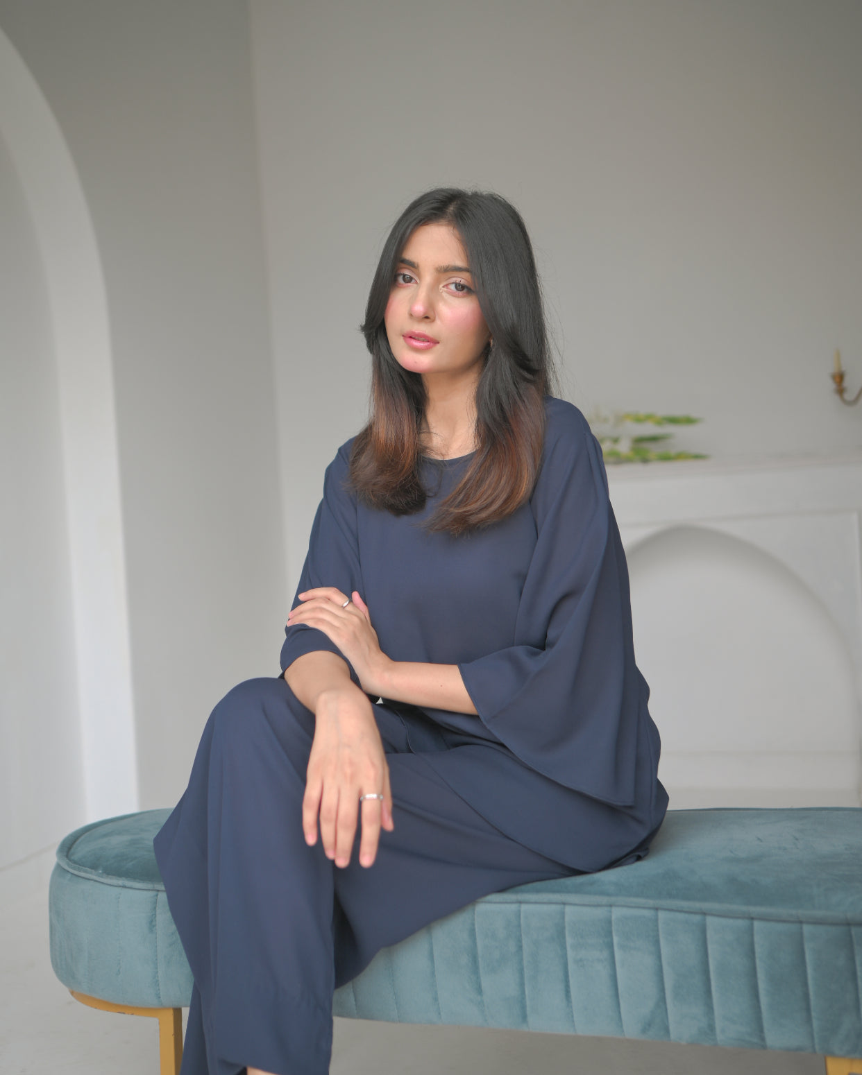 A woman sitting in a navy blue outfit stands next to an arched alcove in a minimalist white room.