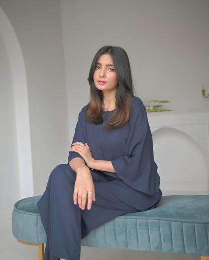 A woman sitting in a navy blue outfit stands next to an arched alcove in a minimalist white room.