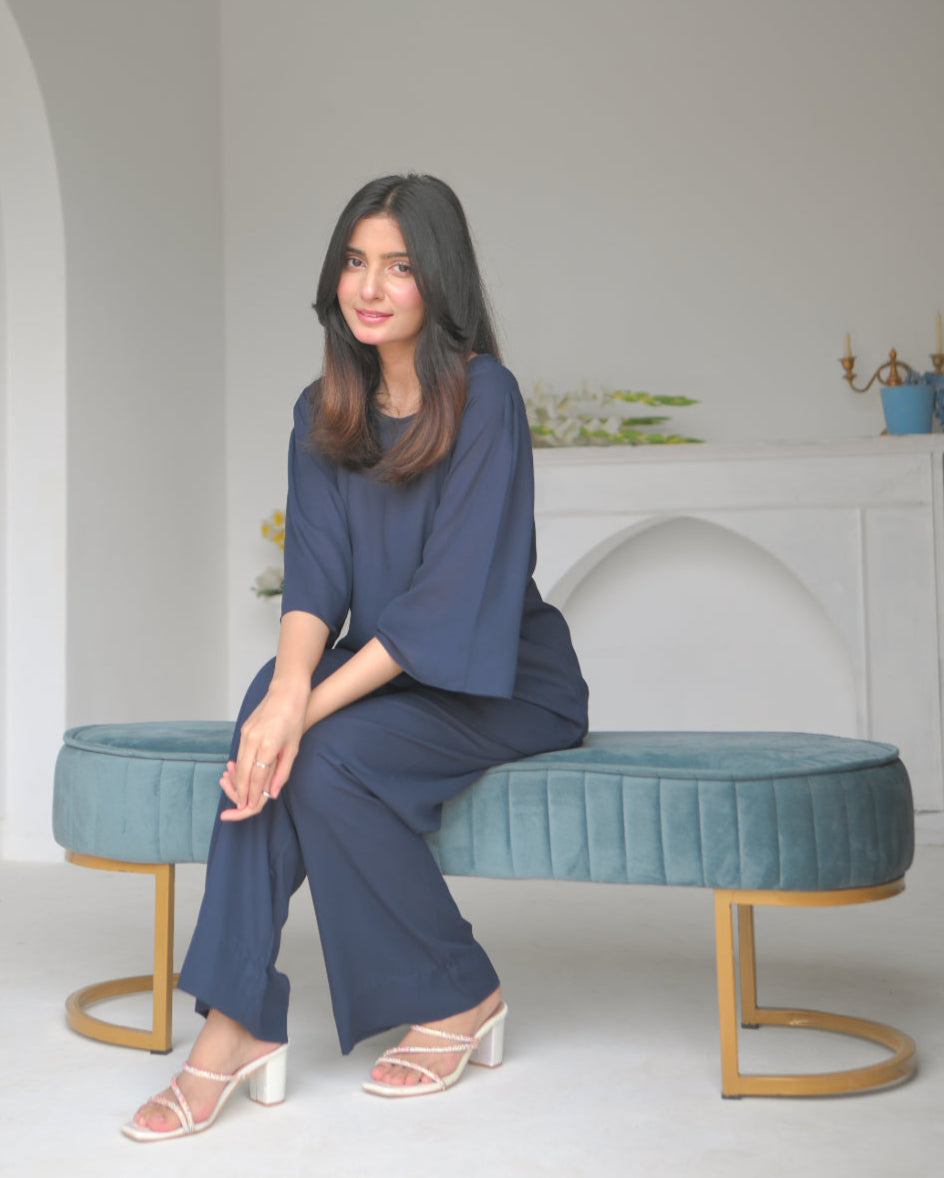 A woman sitting in a navy blue outfit next to an arched alcove in a minimalist white room.