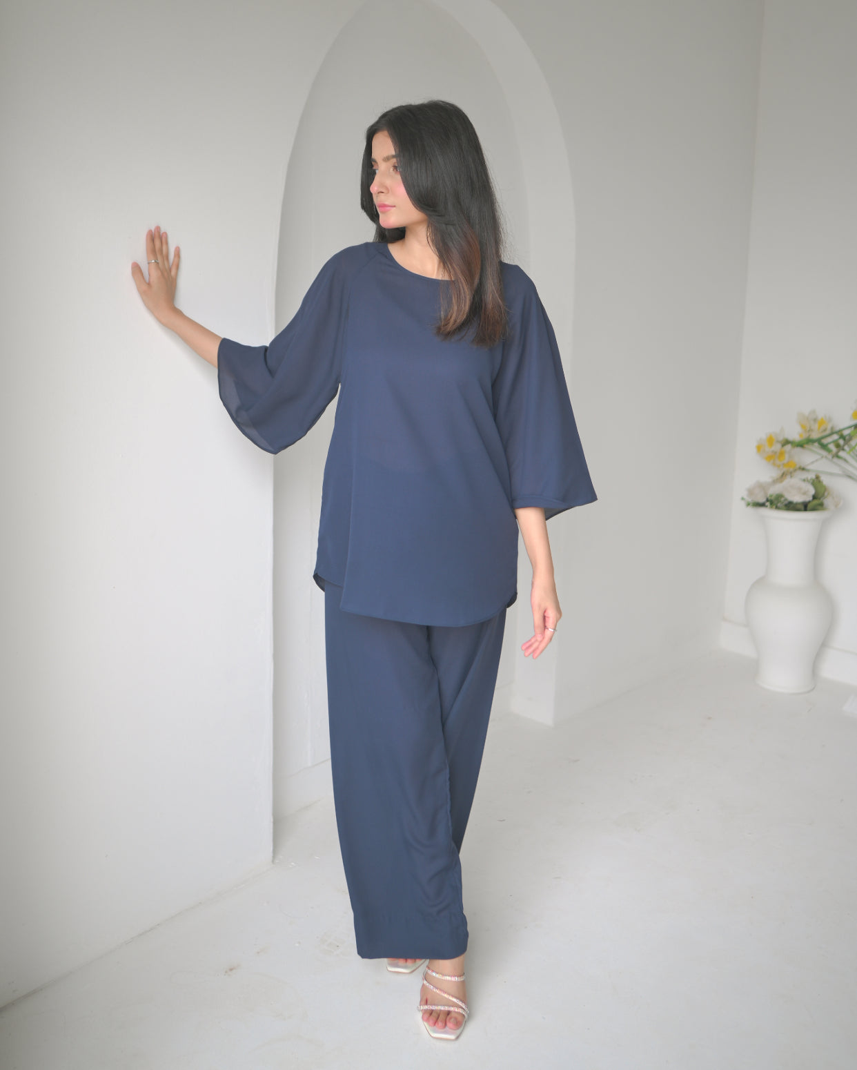 A woman in a navy blue outfit stands next to an arched alcove in a minimalist white room.