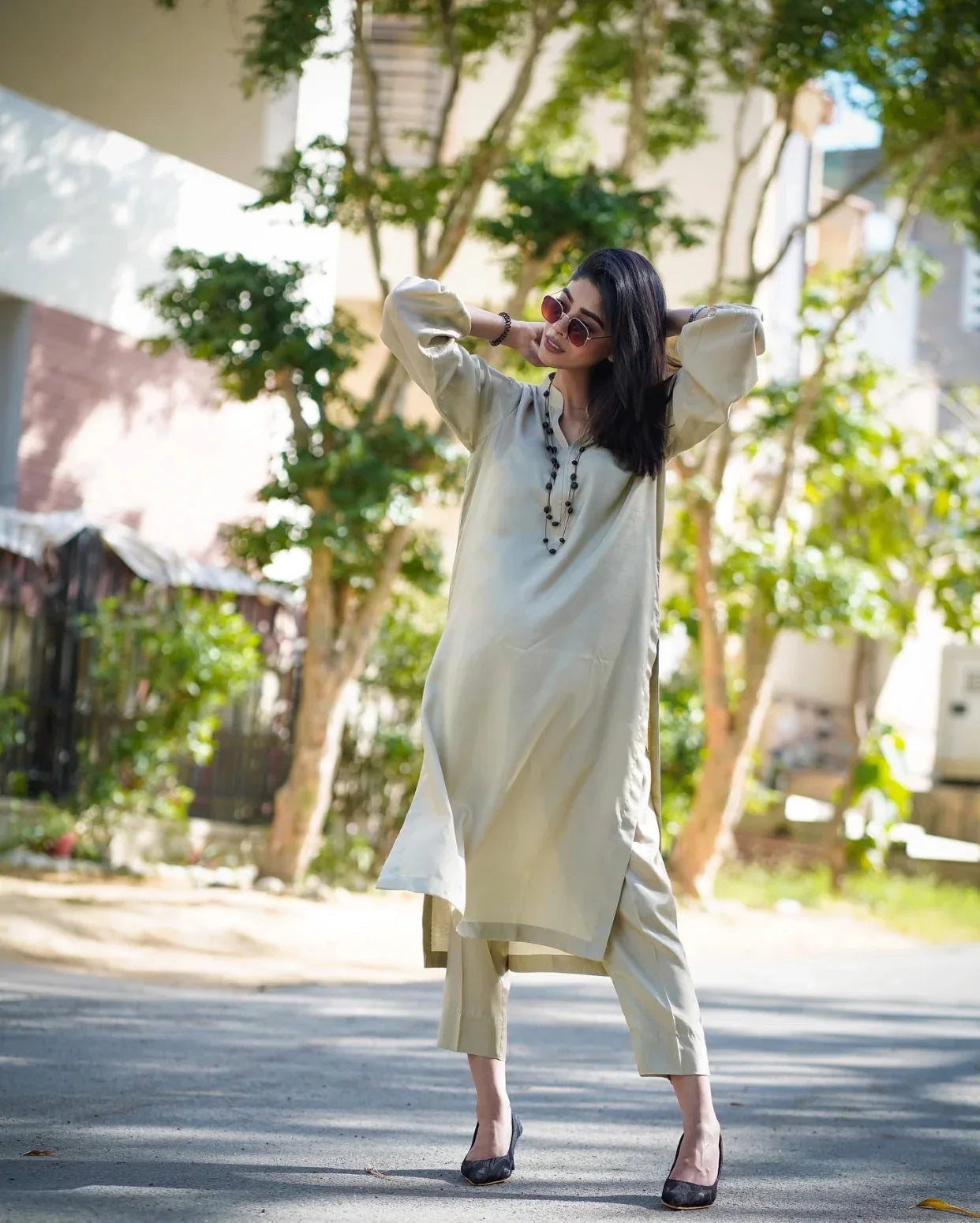 Woman in a beige outfit standing outdoors with trees and buildings in the background