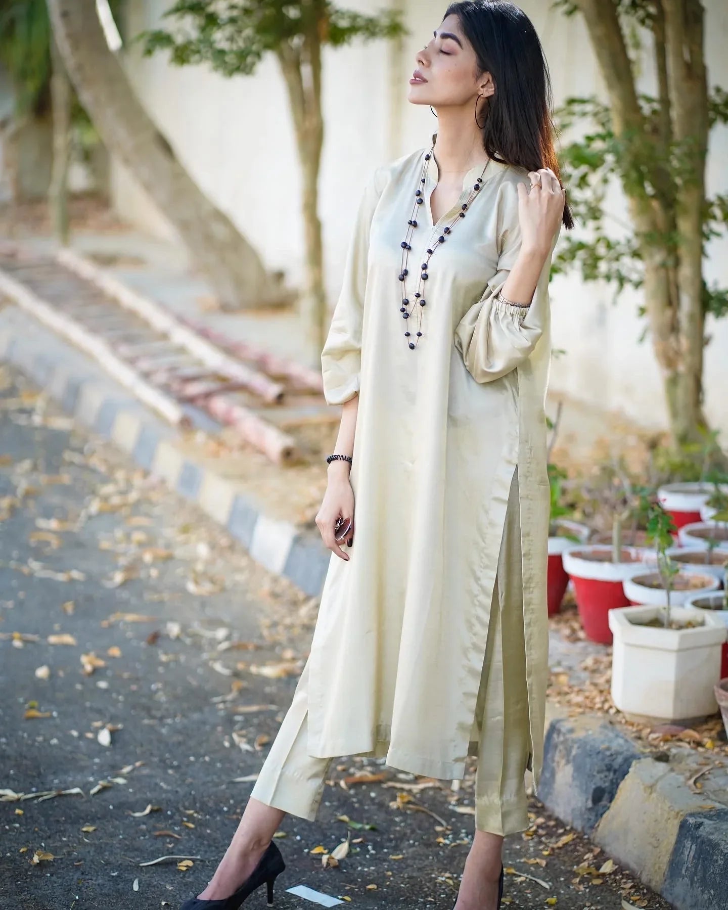 A women wearing a beige dress kurta  standing outdoors with a confident pose. Background is leafy and sunlit.