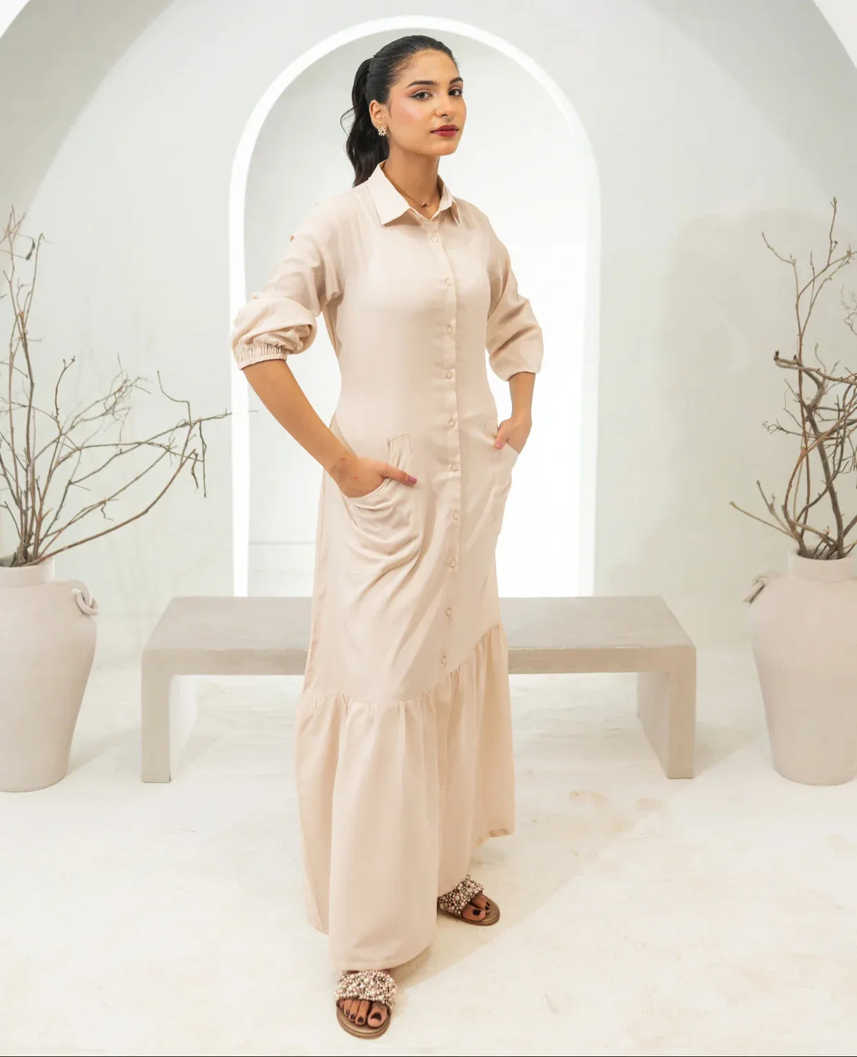 Woman in a long, fawn color dress standing in a minimalistic room with white walls and a bench.