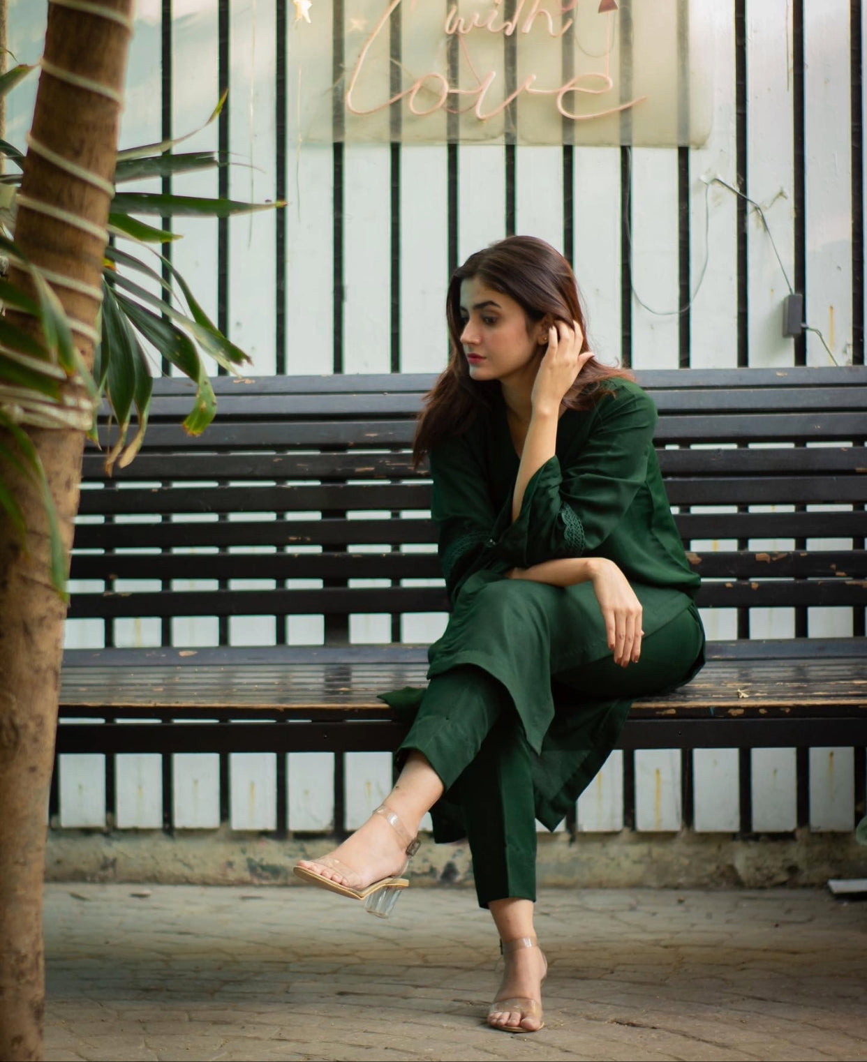 Woman in a bottle green outfit sitting on a bench with plants and a decorative sign in the background