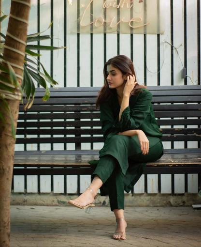 Woman in a bottle green outfit sitting on a bench with plants and a decorative sign in the background