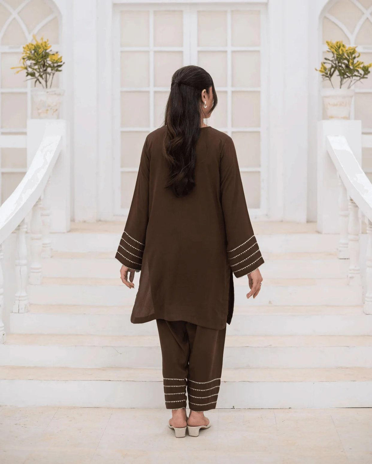 A woman viewed from behind wearing a brown co ord set with silver and gold laces on the cuffs and hems, standing on white stone steps in front of a white arched building.
