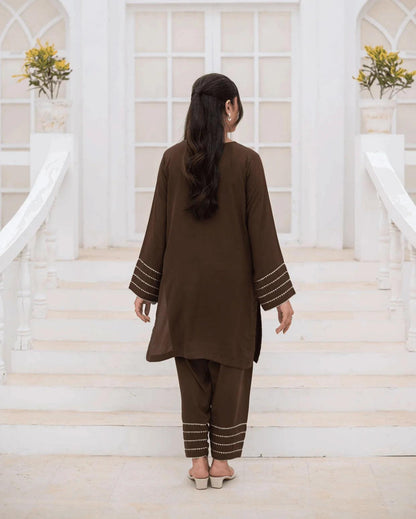 A woman viewed from behind wearing a brown co ord set with silver and gold laces on the cuffs and hems, standing on white stone steps in front of a white arched building.
