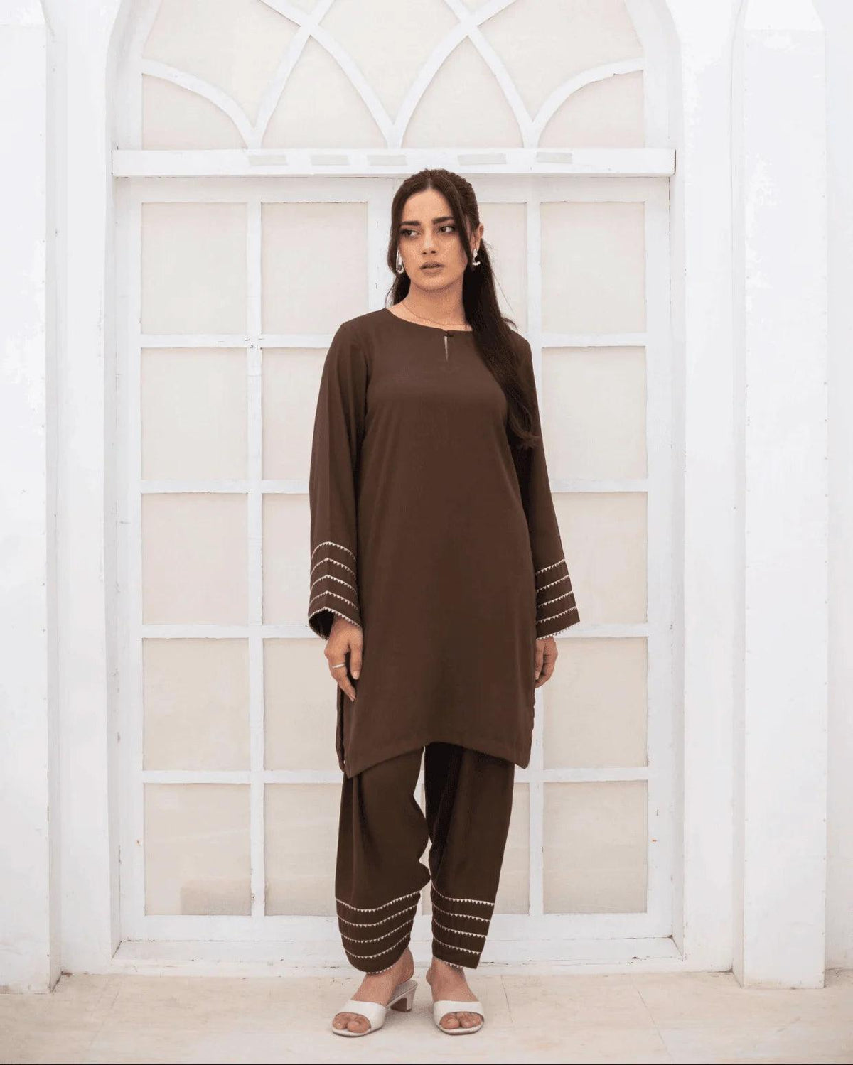 A woman wearing a brown co ord set with silver and gold lace on sleeve and hems, standing in front of a white arched window.