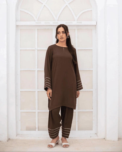 A woman wearing a brown co ord set with silver and gold lace on sleeve and hems, standing in front of a white arched window.