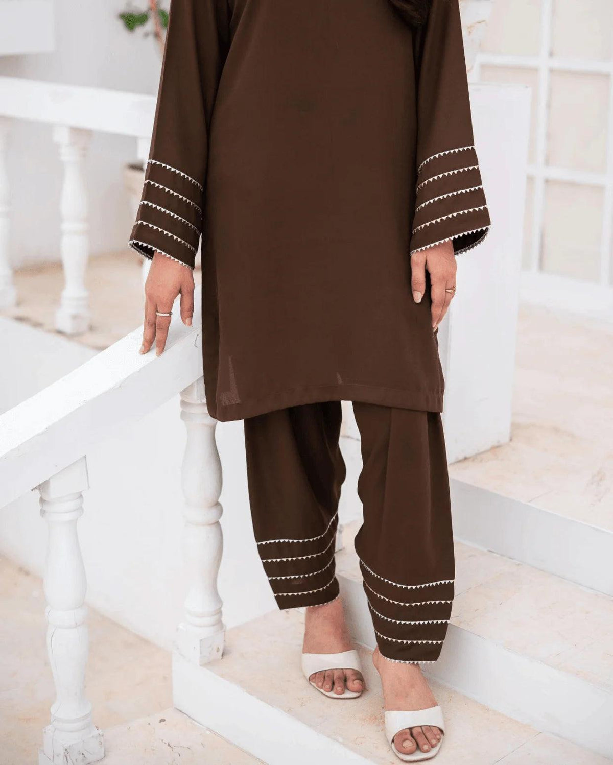 A woman wearing a brown co ord set with silver and gold lace near the cuffs, standing on white steps in a bright indoor setting.