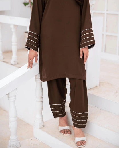 A woman wearing a brown co ord set with silver and gold lace near the cuffs, standing on white steps in a bright indoor setting.