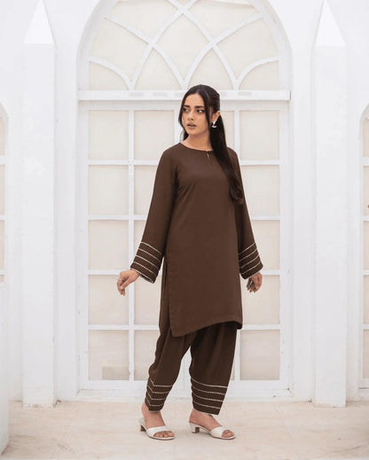 A woman modeling a brown co ord set with silver and gold laces on cuffs and hems, standing in front of a white arched window.