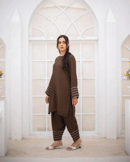 A woman modeling a brown co ord set with silver and gold lace on sleeve and hems, standing in front of a white arched window.