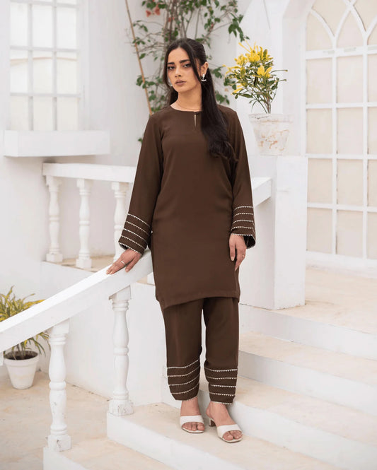 A woman modeling a brown co ord set with silver and gold lace on the cuffs and hems, standing on white steps in a bright, modern white courtyard.