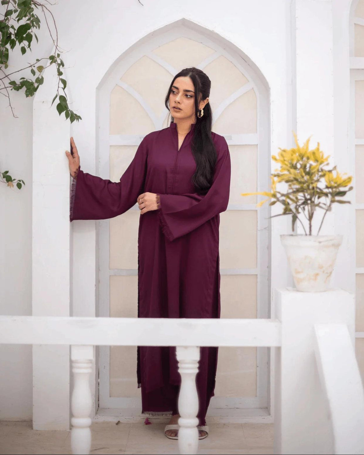 A woman wearing a long-sleeved, burgundy co ord set, standing in a white arched doorway with decorative plants on either side.