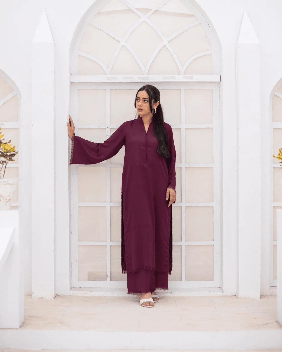A woman wearing a long-sleeved, co ord set stands in front of a white arched doorway with multiple panes, holding onto the white pillar on her left.