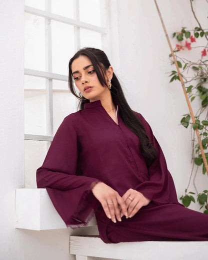 A woman wearing a deep burgundy long-sleeve co ord set, sitting on a white bench in a bright, modern room with a window and climbing green vines.