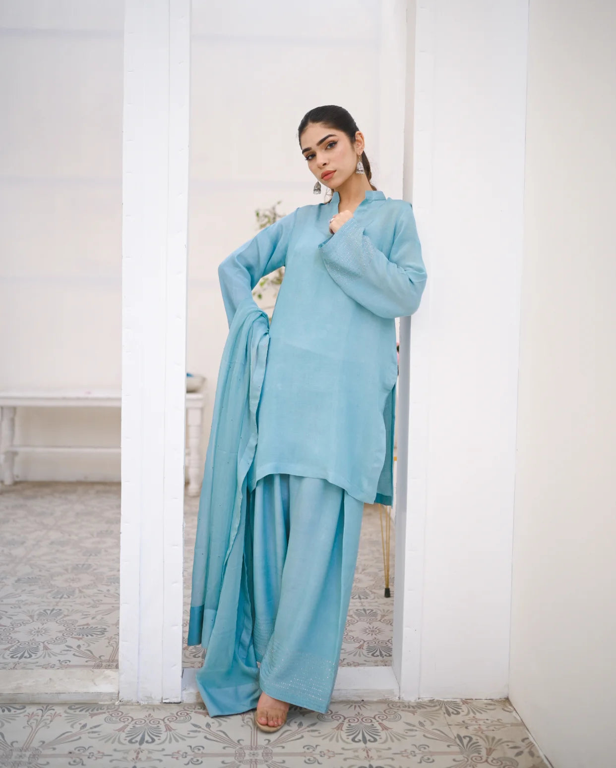 A woman wearing a Sky Blue three piece rawsilk dress which includes a shirt and farshi shalwar with chiffon dupatta. The dress is embellished with stone details.