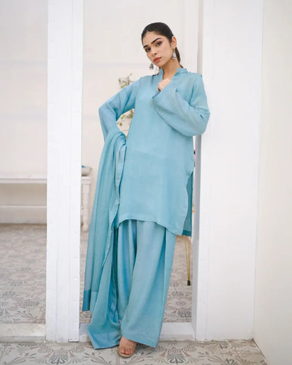 A woman wearing a Sky Blue three piece rawsilk dress which includes a farshi shalwar kameez with chiffon dupatta. The dress is embellished with stone details.
