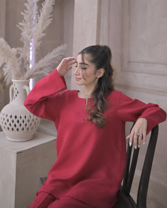 A woman in a deep pink dress sitting on a vintage wooden chair against a beige, textured wall. She has a calm expression.