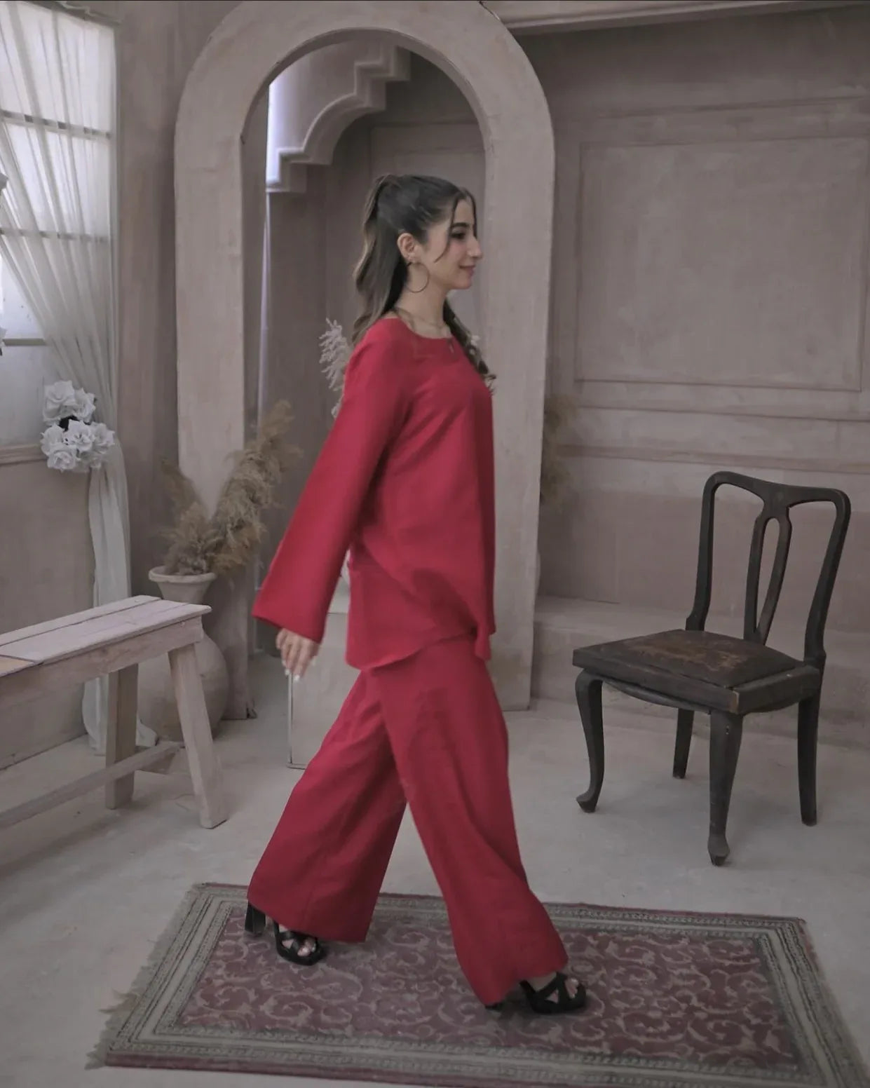 A woman in a deep pink outfit against a beige, textured wall. She has a calm expression and wears black heels.