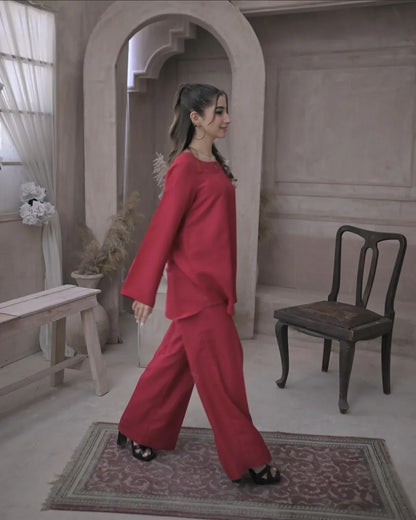 A woman in a deep pink outfit against a beige, textured wall. She has a calm expression and wears black heels.