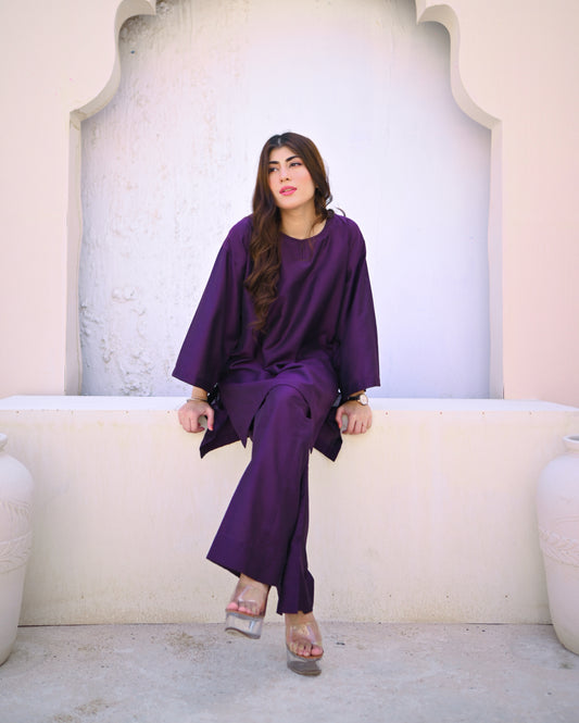 Woman in a purple outfit sitting on a white ledge with a light-colored wall behind her.