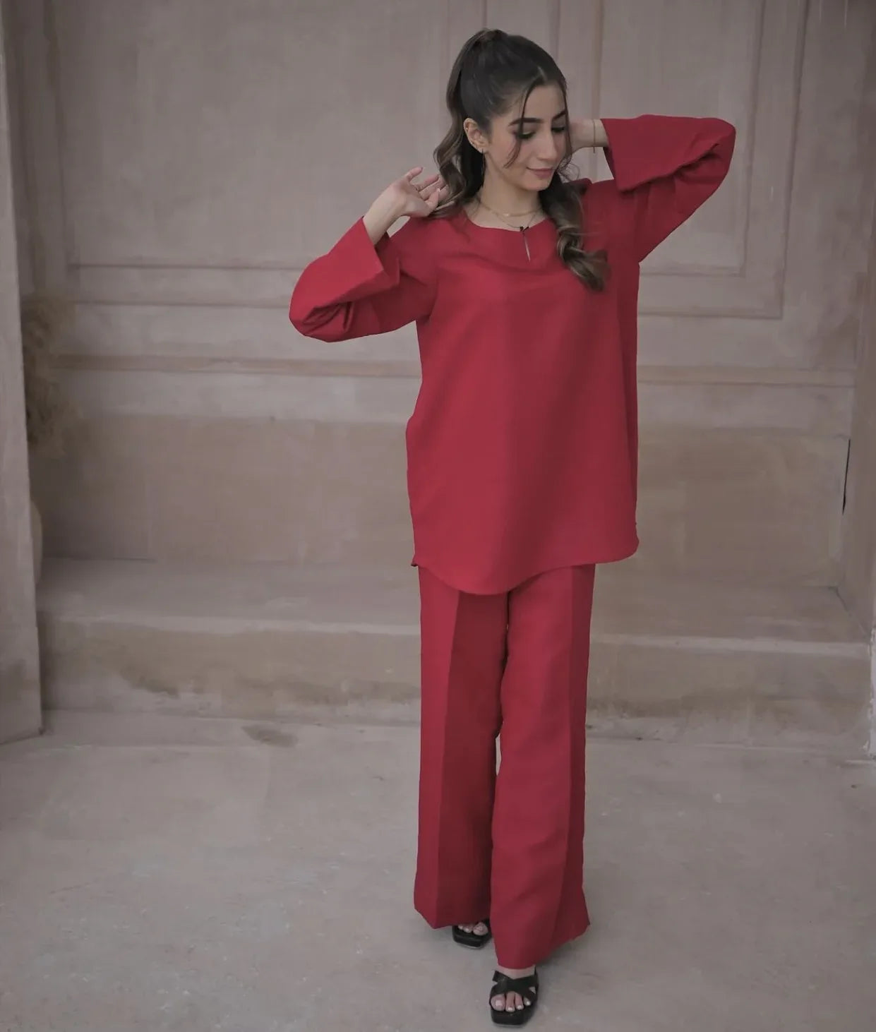 A woman in a deep pink outfit standing against a beige, textured wall. She has a calm expression and wears black sandals.