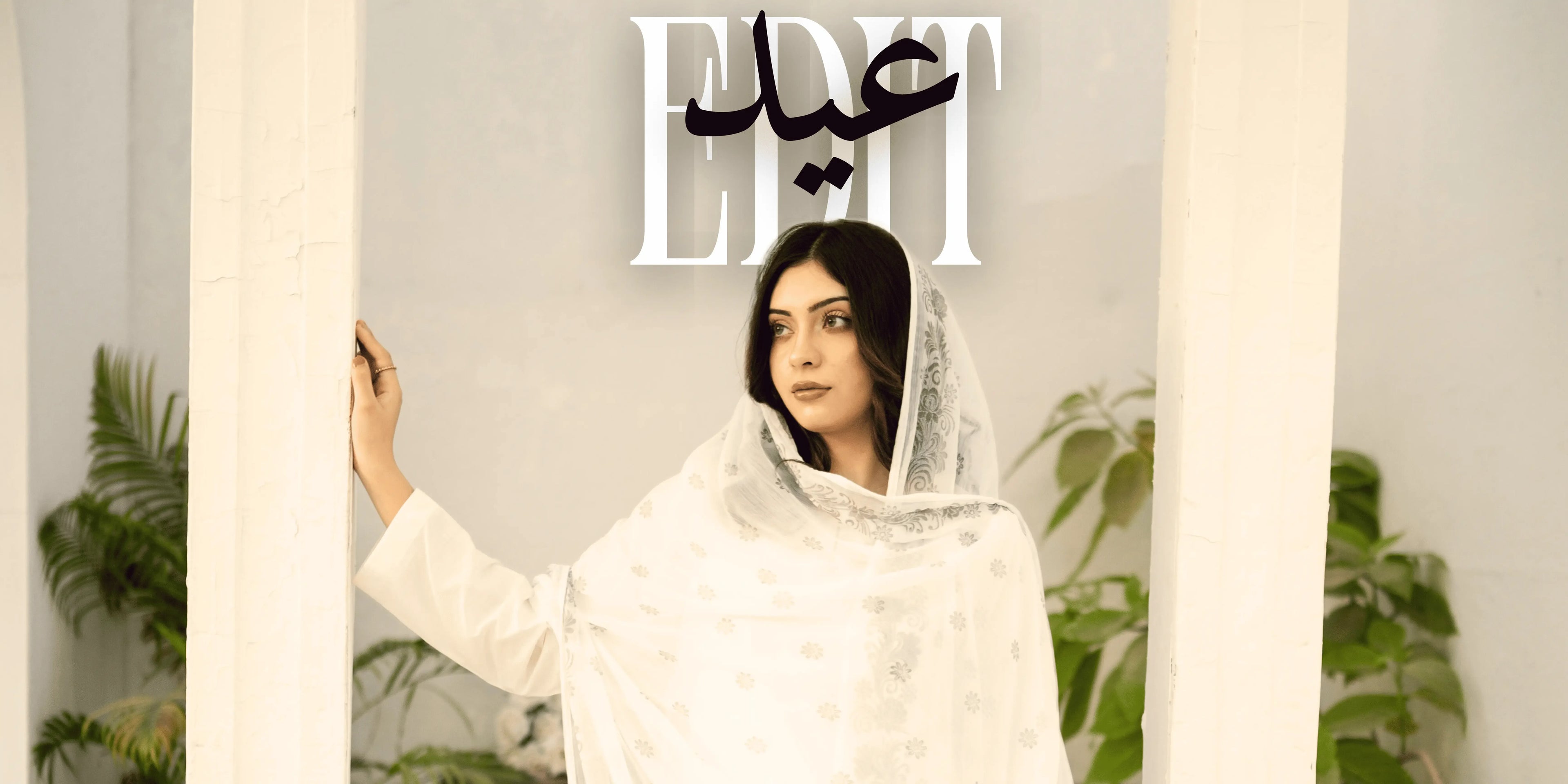 Woman in a white outfit with a headscarf, standing indoors with plants in the background.