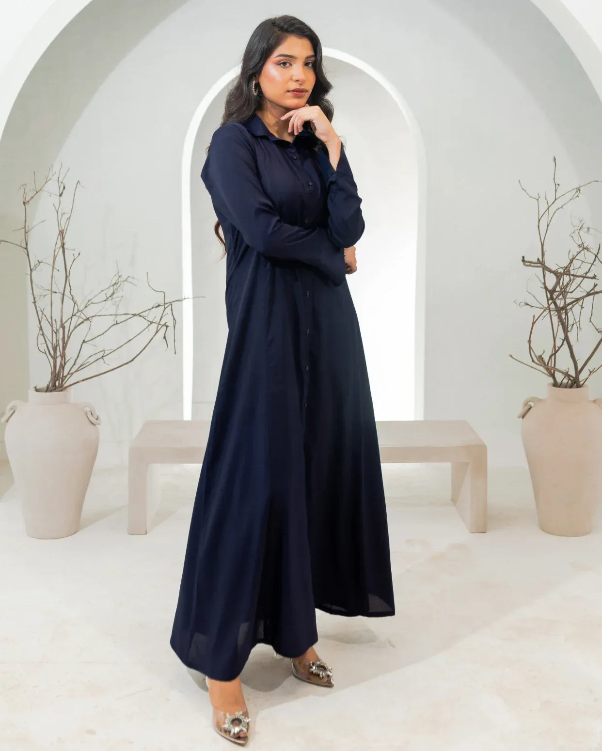 Woman wearing a long navy blue dress in a minimalistic setting with white arches and beige vases.