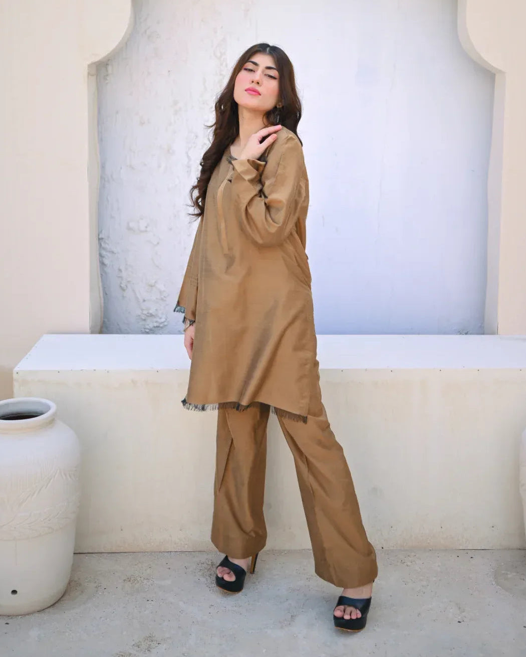 Woman in a beige outfit standing in front of an arched stone wall.