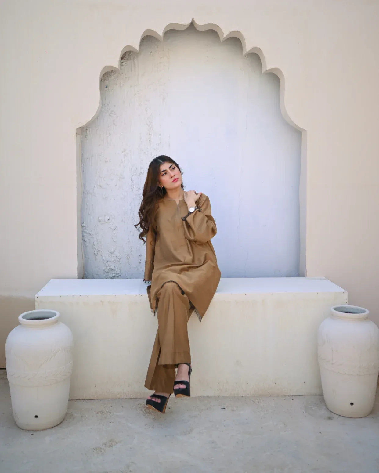 Woman in a beige outfit sitting on a white bench with decorative arch and vases.