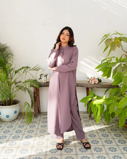 Woman in a long lavender color dress standing in a room with plants and a tiled floor.